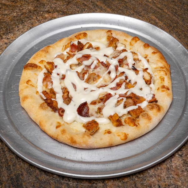 A round pizza with white sauce, chicken, and bacon on a metal pizza pan.