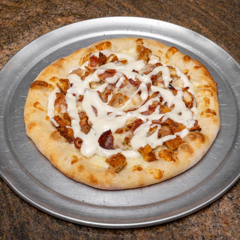 A round pizza with white sauce, chicken, and bacon on a metal pizza pan.