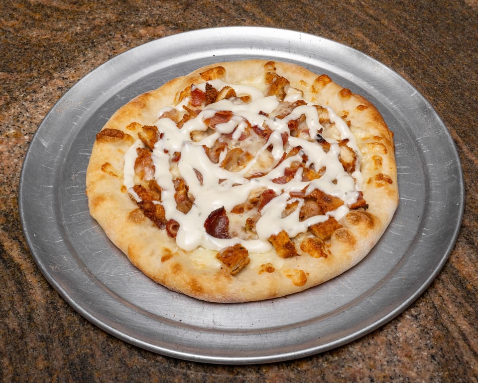 A round pizza with white sauce, chicken, and bacon on a metal pizza pan.