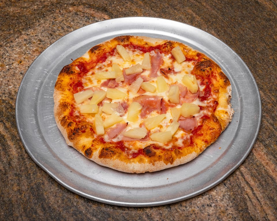 A Hawaiian pizza with ham and pineapple on a metal tray, on a granite countertop.